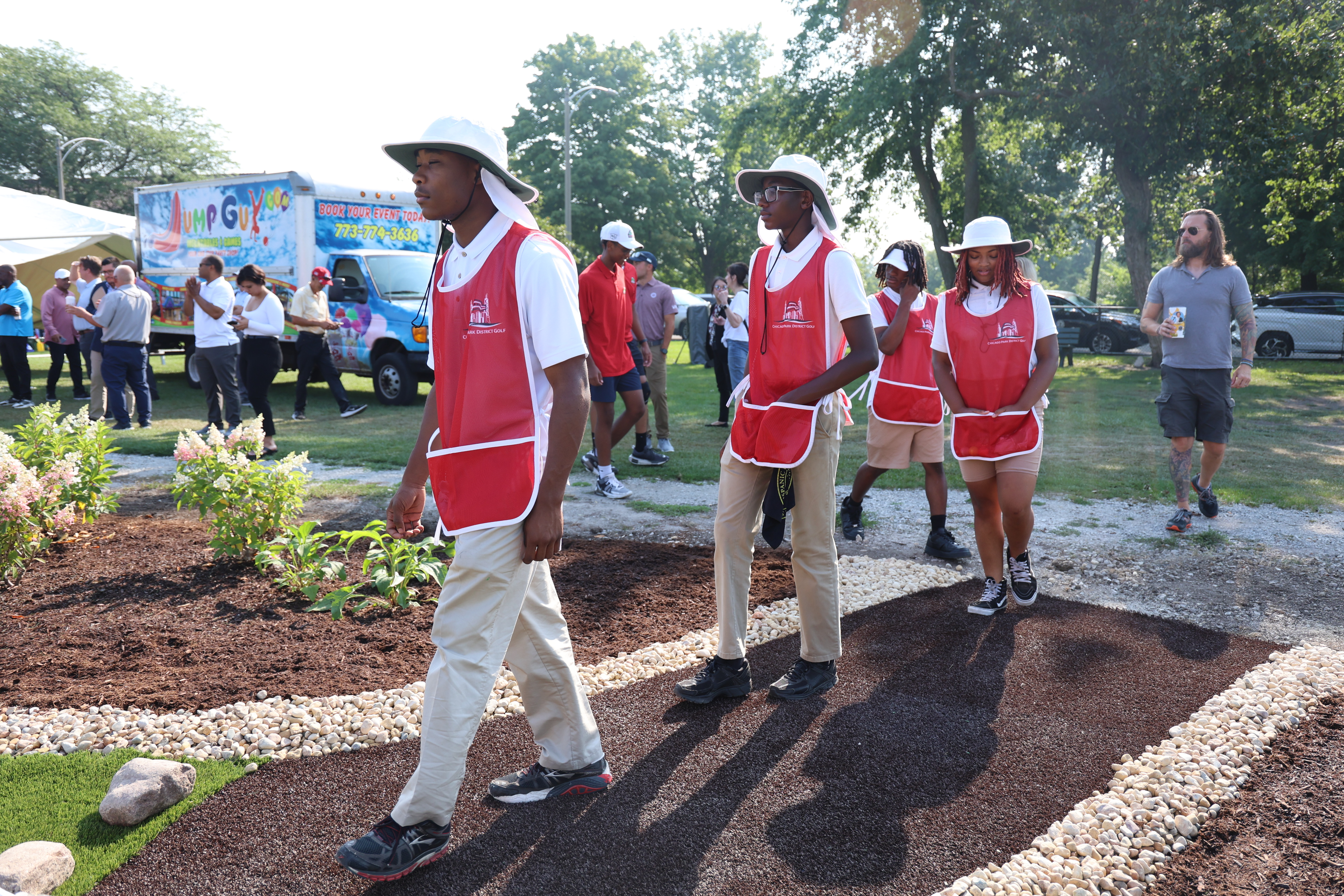 Three young people in red golf caddie vests walk on a path by a landscaped area.